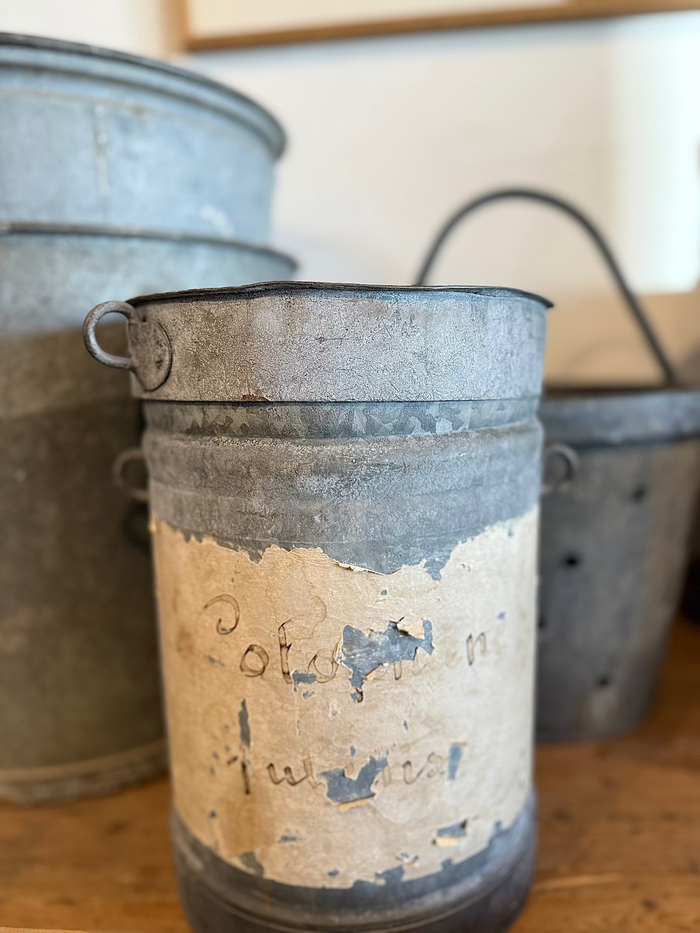 Weathered metal canister with flat lid and side handles; worn blue-and-tan artwork